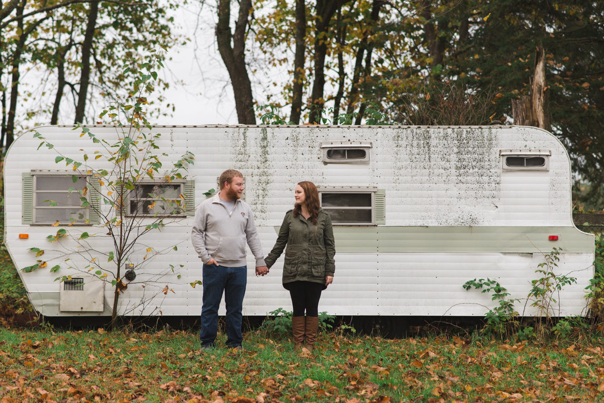 engaged couple standing in front of old RV trailer holding hands