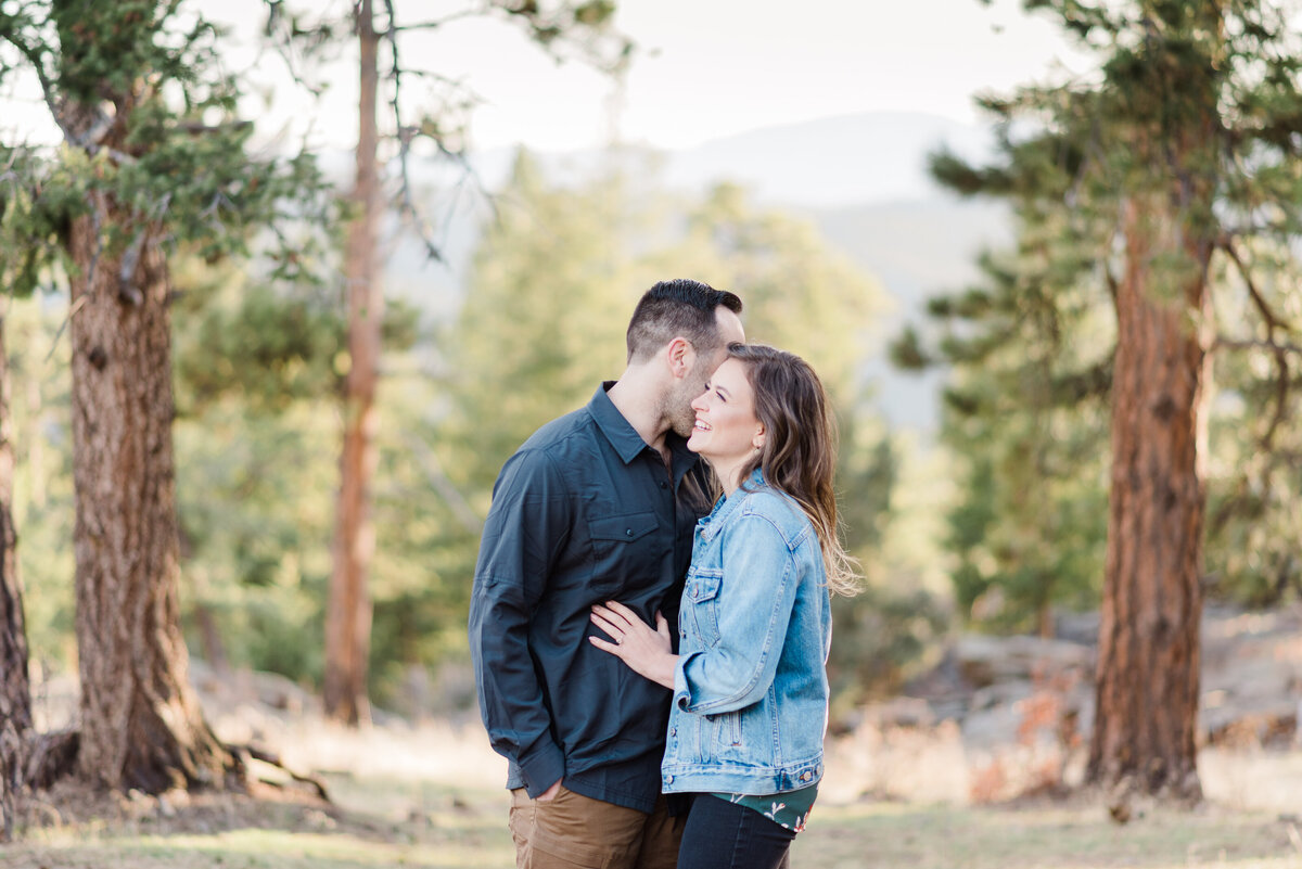 outdoor colorado engagement photos with man and woman embracing each other in the woods with man whispering in woman's ear as she laughs captured by denver engagement photographer