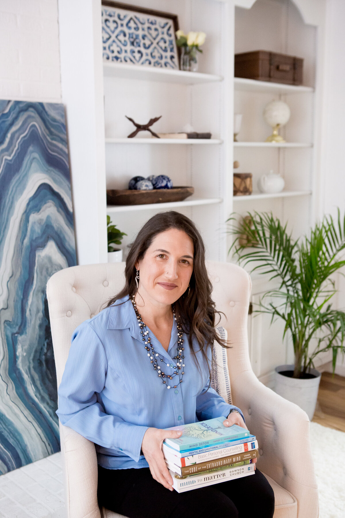 professional young woman in a blue blouse sits in a white wingback chair holding a stack of books in a bright office with a green plant in the background New York branding photographer Milestone Images
