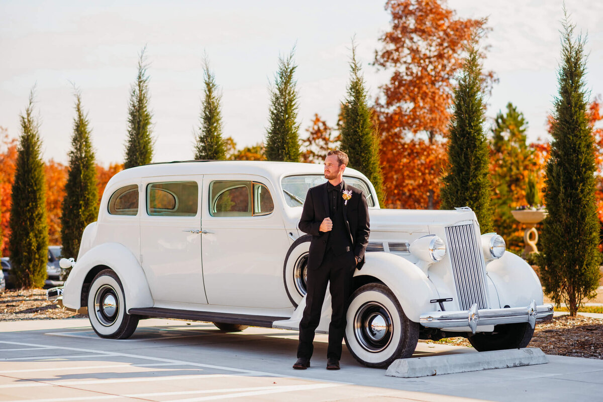 Photo of a groom in a black suit leaning on a white vintage vehicle
