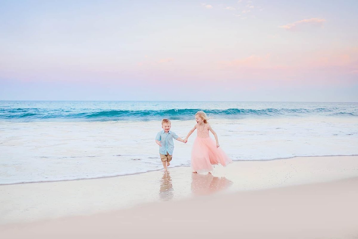 Love + Water family session at sunrise with young girl in pink dress and young boy in blue collared shirts holding hands and running on the beach