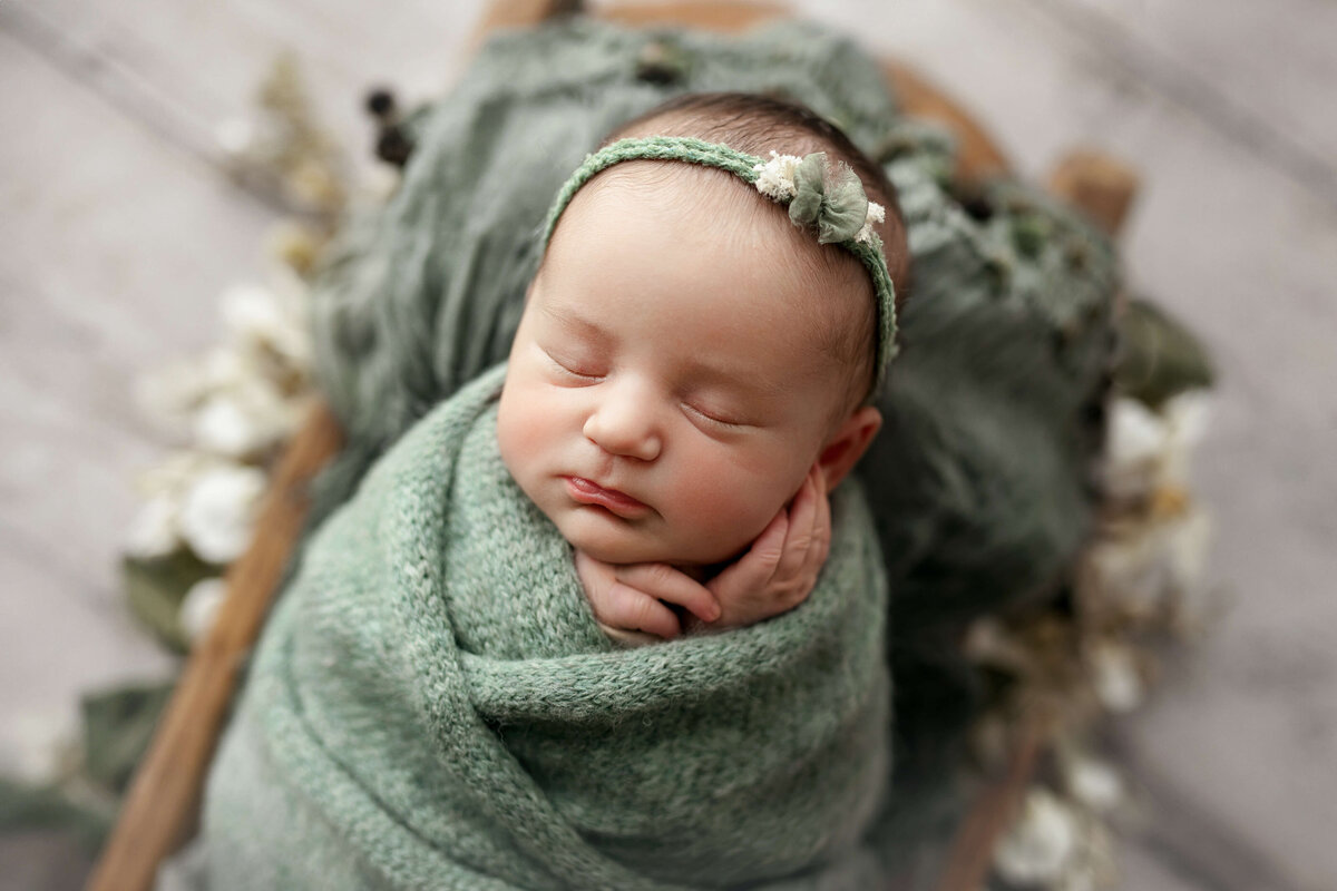 newborn baby birl wrapped in a green knit wrap with a matching headband laying on her lack in a wooden trench bowl prop at a loudoun county va newborn photo session