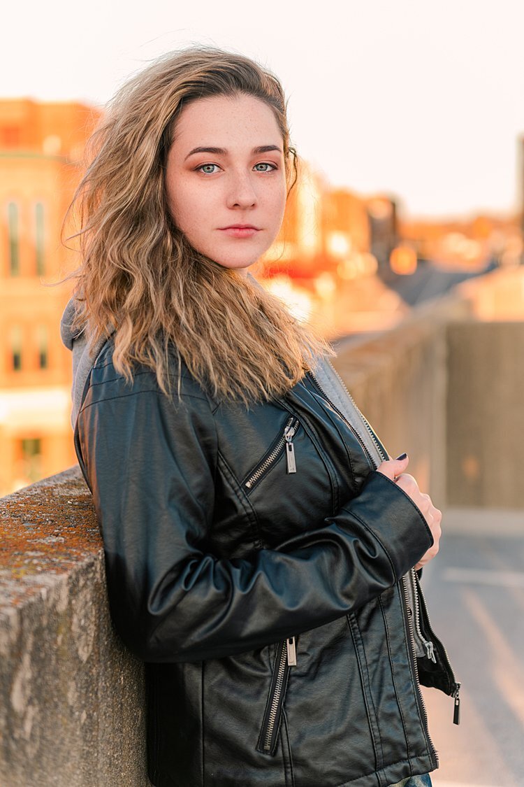 High school senior girl in black leather jacket leaning against wall on roof of parking garage at sunset