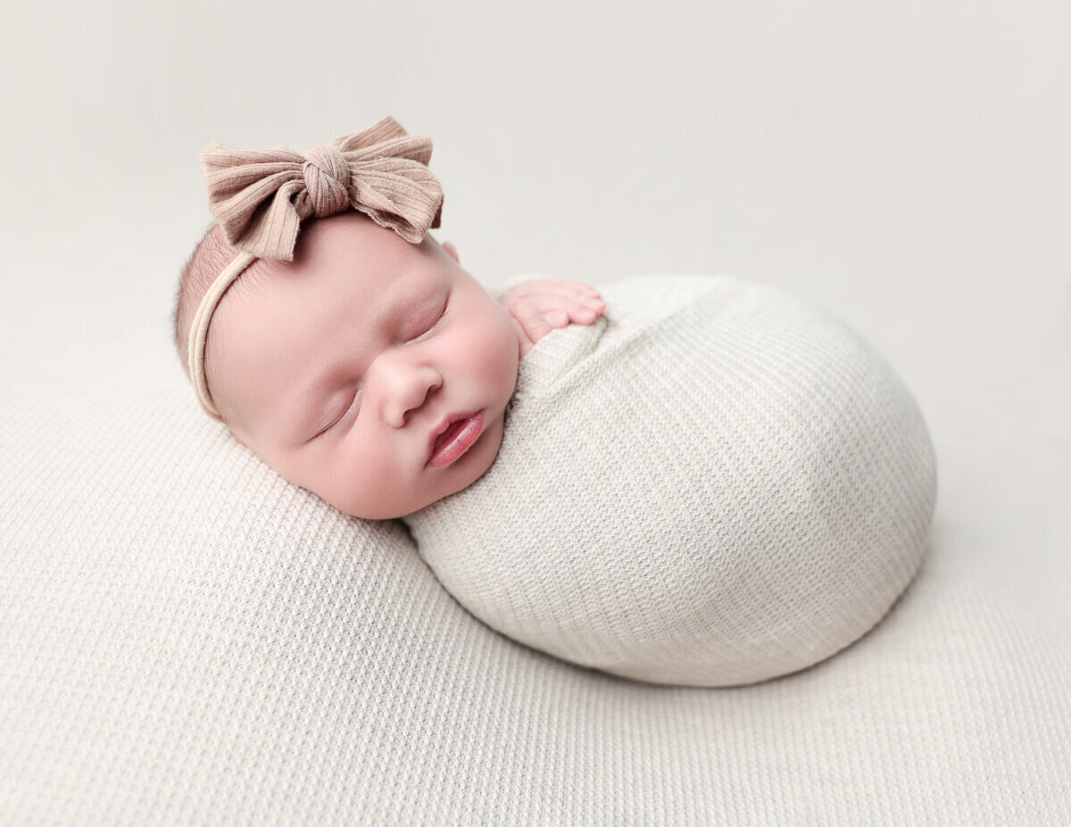 A beautiful newborn girl swaddled in a soft tan wrap, resting peacefully on a matching tan backdrop in a photography studio located in Rochester, NY.