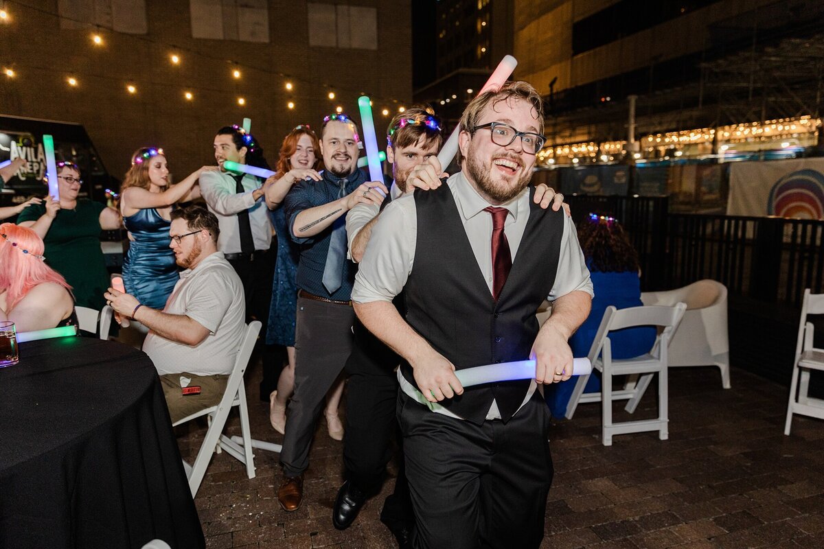 A groom leading a conga line of guests during his wedding reception at the Pegasus City Brewery in Dallas, Texas. The groom is wearing a suit (minus the jacket), and he and the guests are all holding light up foam sticks.