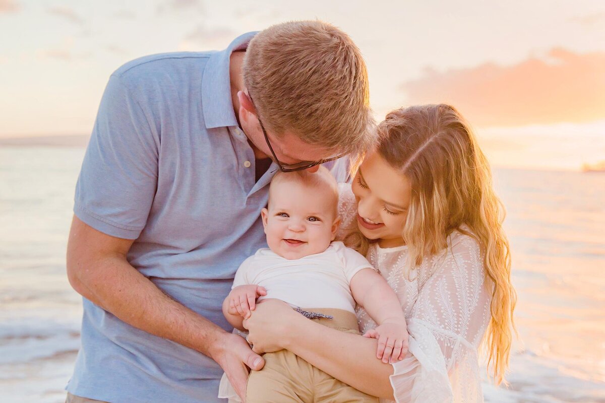 Young family cuddling their smiling baby backlit by the setting sun on Maui during a photography session with Love + Water