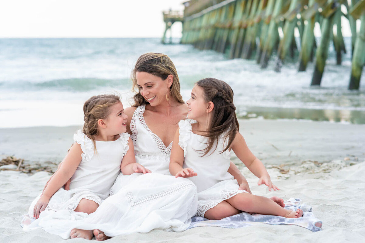 At the beach, next to a pier, a mom and her two daughter dressed in all white sit staring lovingly at each other on the sand.