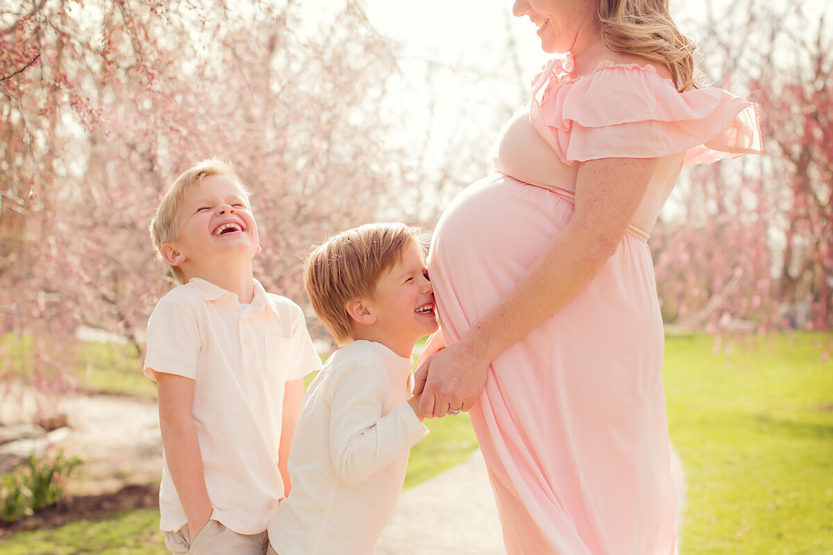 Mom is outside in the spring in a flowing pink dress, cherry blossoms behind her, with her young boys giggling.