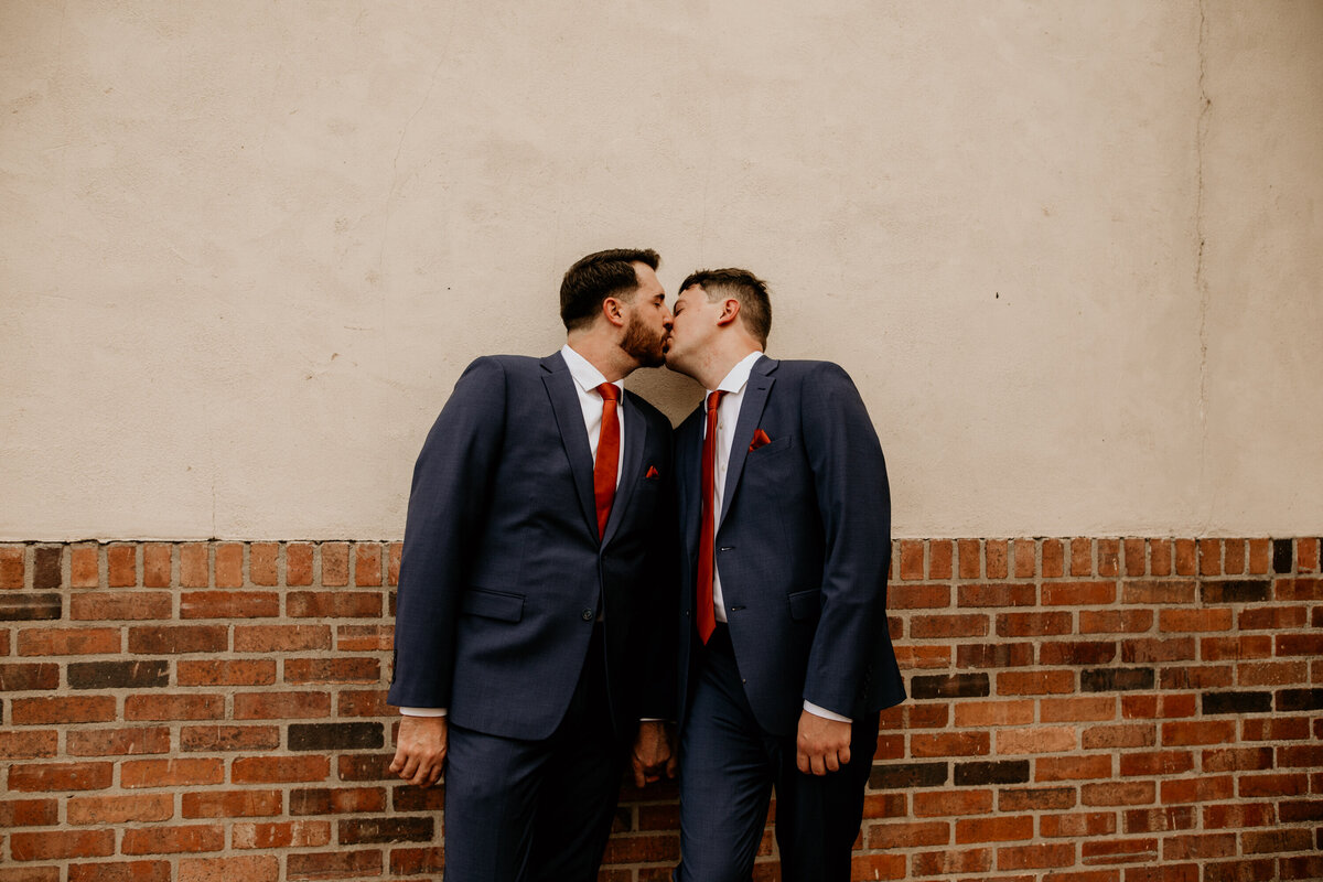 two grooms standing against a building holding hands in downtown Albuquerque kissing