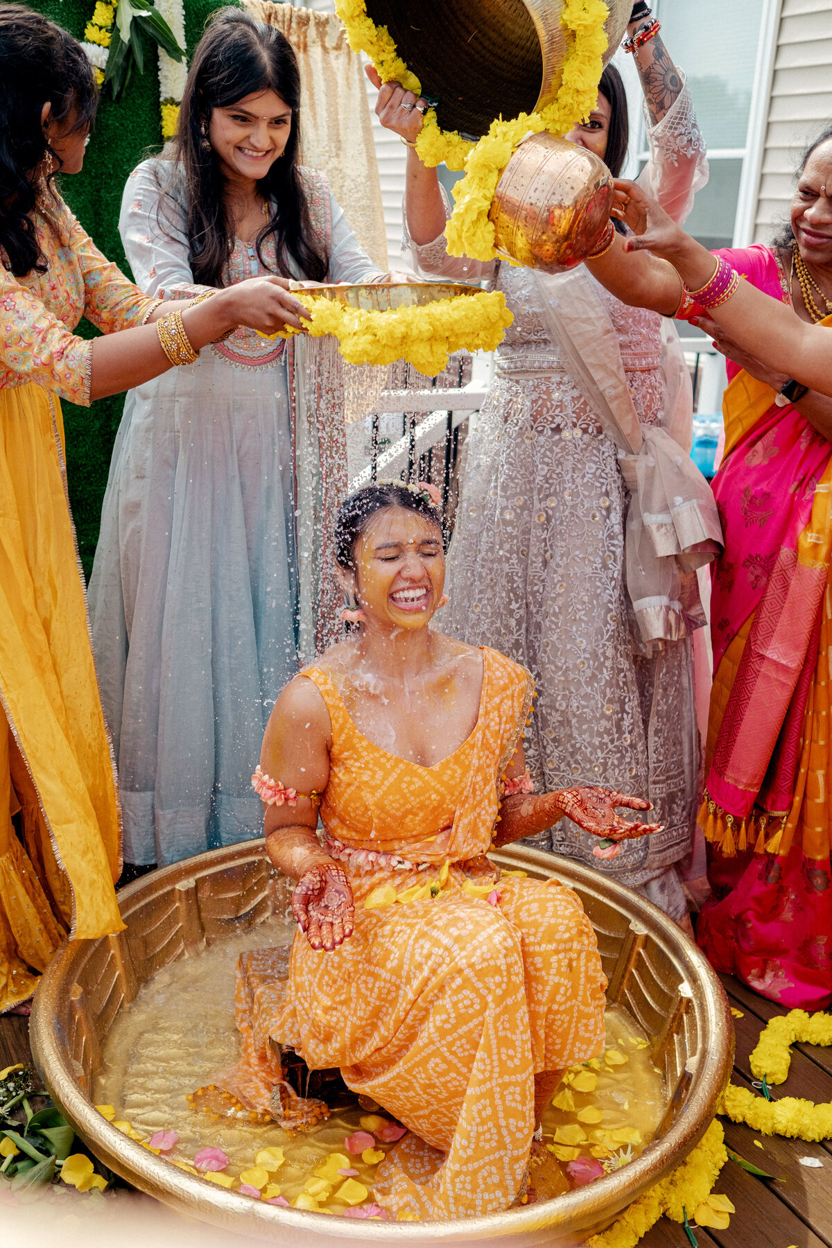 A bride is being showered after her haldi ceremony.  She is wearing a yellow gown  and is smiling while water is sprinkled on her