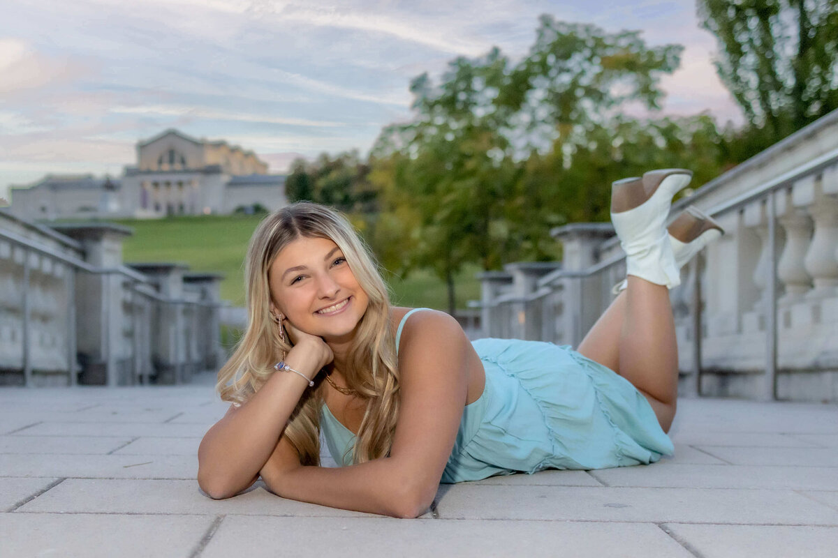 a blonde haired high school senior girl  p\laying on her stomach and propped up with her arms and legs pulled up  behind her and feet crossed on a bridge in a park