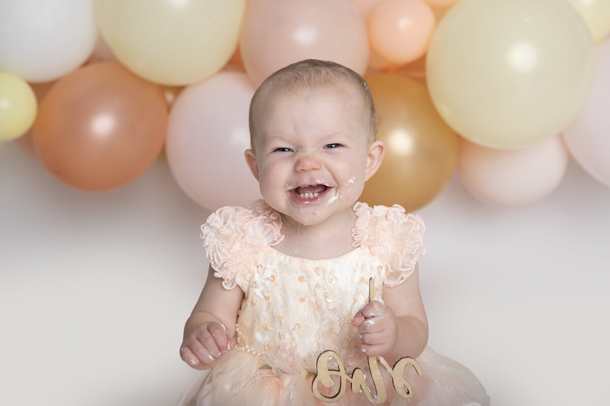 First birthday baby girl eating a white cake with a pink yellow and peach balloon garland