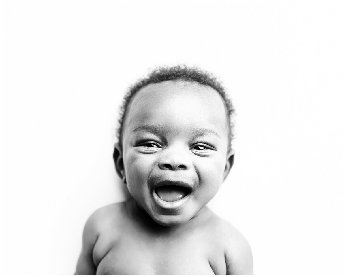 Beautiful baby boy laying on his back on white backdrop looking at the camera with a huge smile on his face.