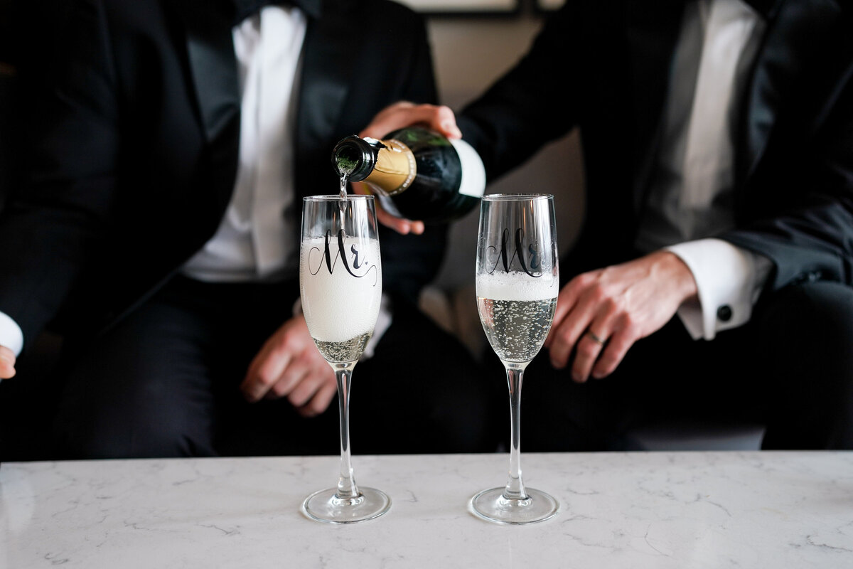 Two grooms in tuxedos sit on a couch on wedding day at Salvage One in Chicago and pour a glass of champagne to celebrate.