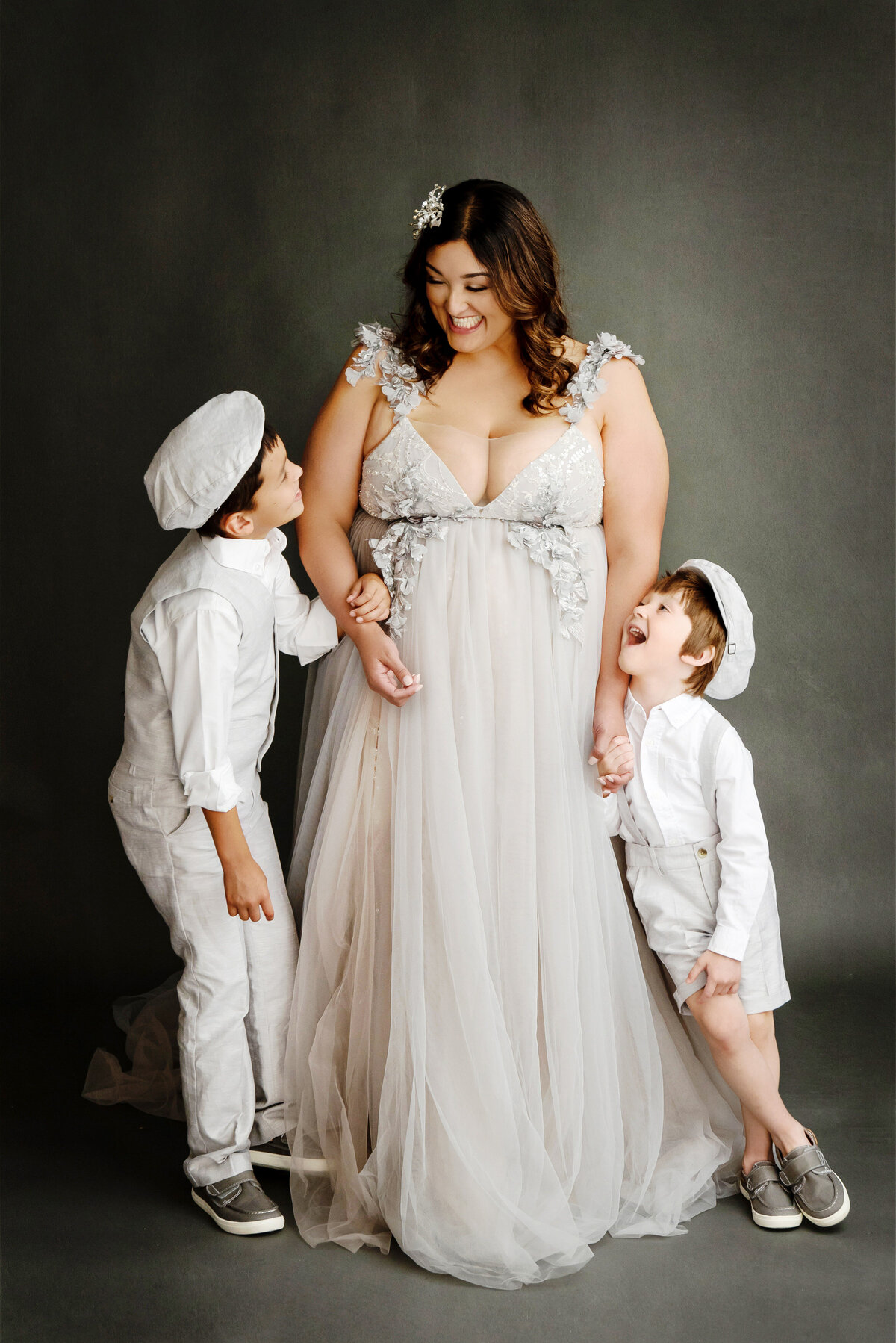 st-louis-motherhood-photographer-mother-in-silver-tulle-gown-with-floral-details-holding-sons-hands-on-either-side-wearing-white-newsboy-outfits-similing