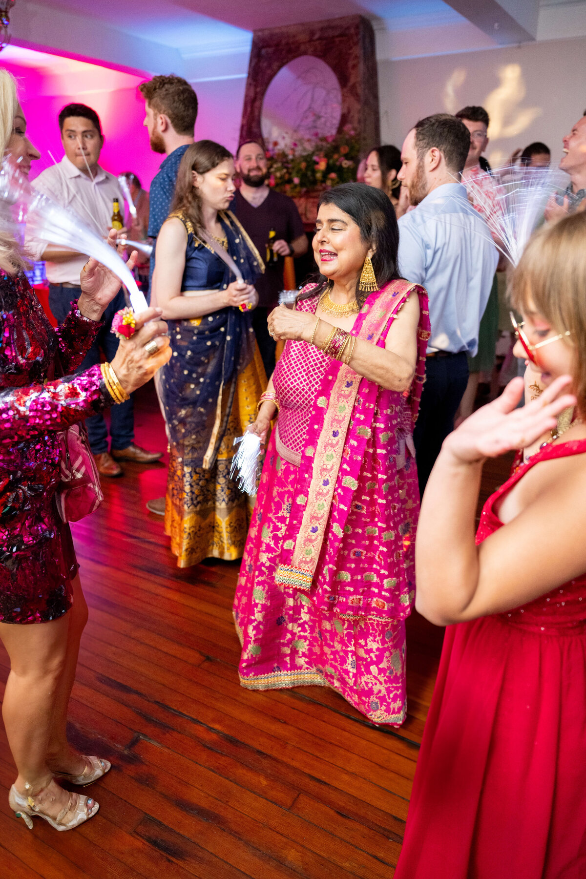 A group of people dancing at a party. The central figure, a woman in a vibrant pink traditional outfit, is smiling and dancing. Others around her are engaged in conversation and enjoying the lively atmosphere. The room is warmly lit with colorful lights.