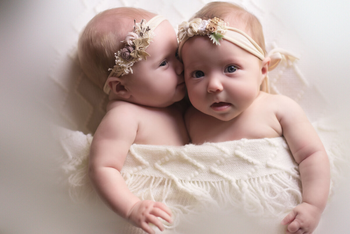 Twin newborn girls posed together one is laying on her back and the other is laying on her side and looks like she is whispering into her sister's ear. They are covered in beautiful textured cream color cloth and wearing headbands with flowers and leaves.