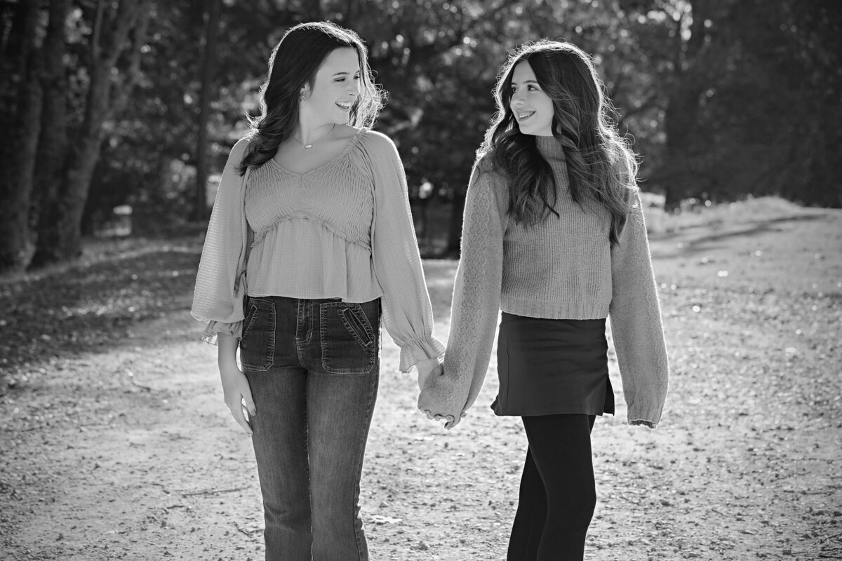 HS senior and younger sister back to back smiling on tree‑lined gravel road in autumn at Anne Springs Close Greenway in black and white