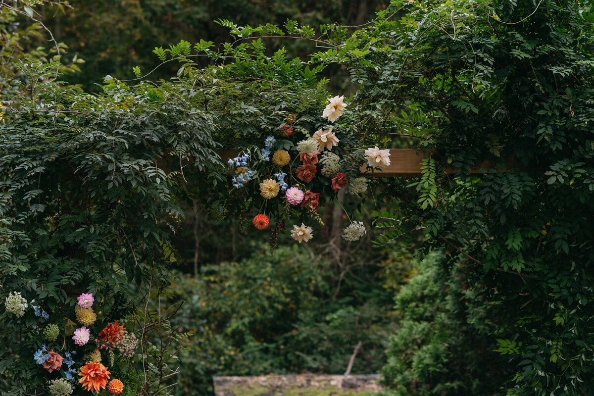 Full view of a garden-style floral arch installation blending into existing greenery with colorful dahlias, roses, and textural blooms for an outdoor ceremony at The Nest in Northwest Arkansas.
