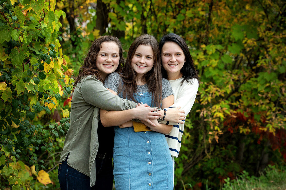 Three young adult sisters hugging with bushes mixed with green and fall colors behind them.