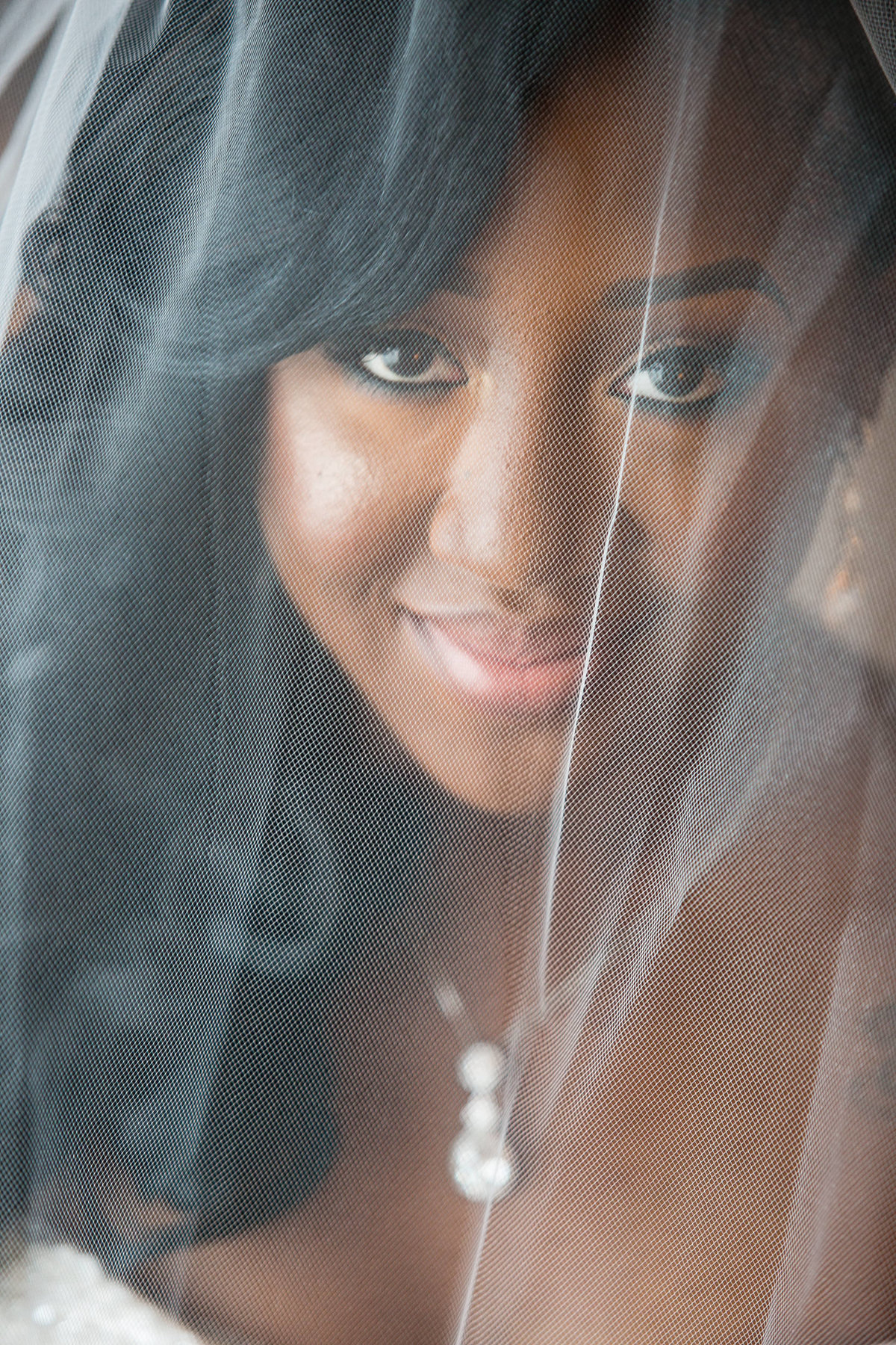 Stunning, close up bridal portrait of Black woman in her wedding dress, veil covering her face, smiling at the camera.