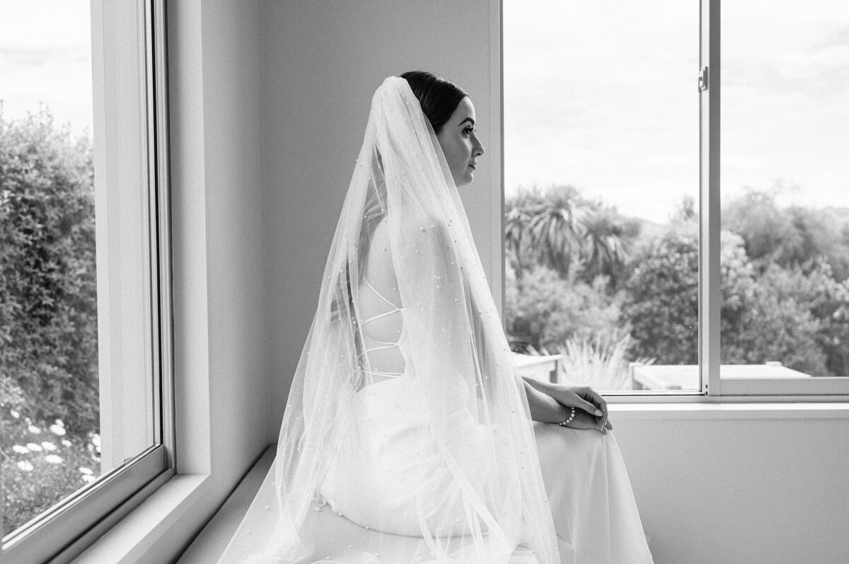 black and white side on portrait of bride with pearl veil and grace loves lace dress sitting by window on wedding day at chirstchurch