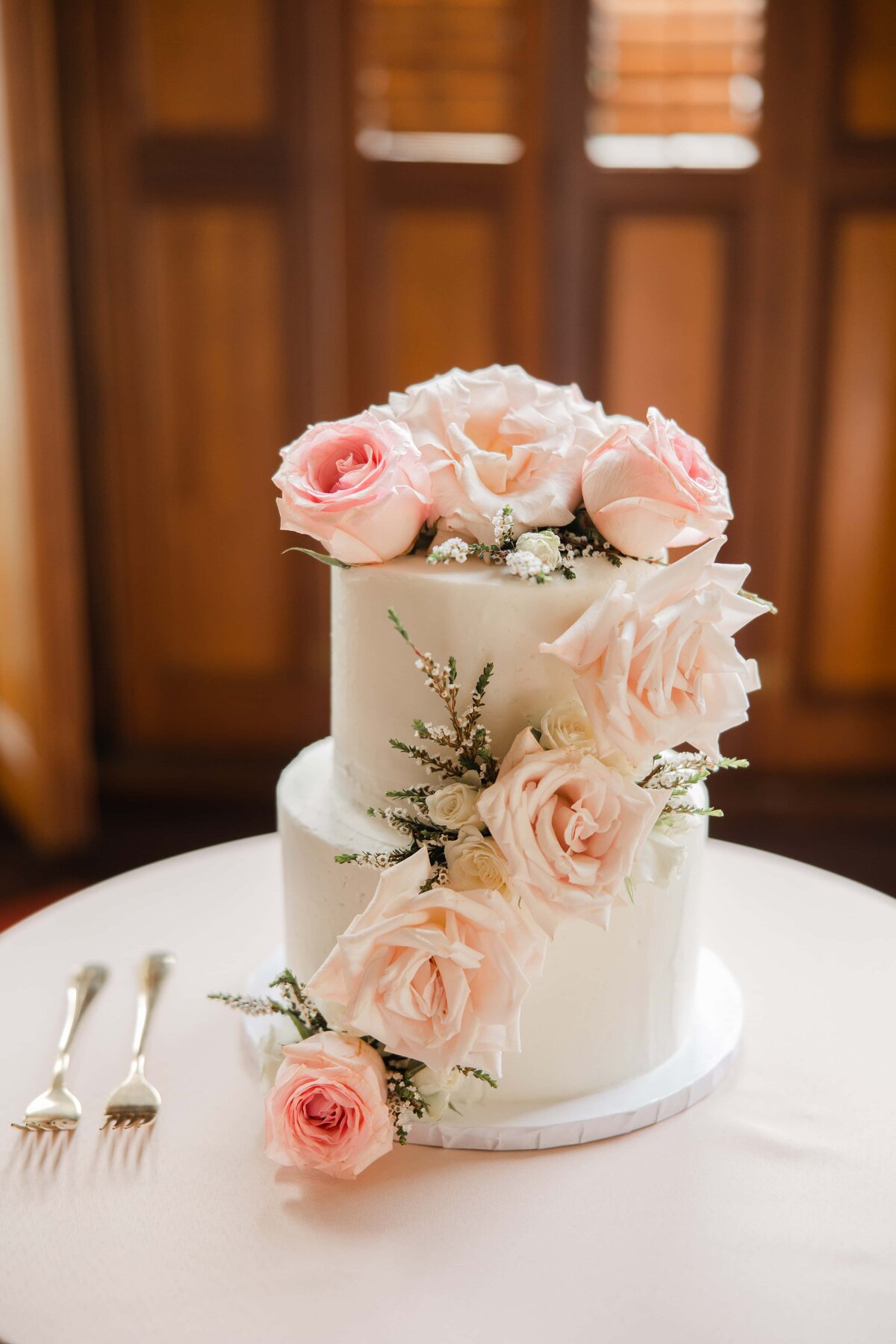 Two-tiered white wedding cake adorned with pink roses and baby's breath on a table at a Park Farm Winery wedding, with cake servers nearby.