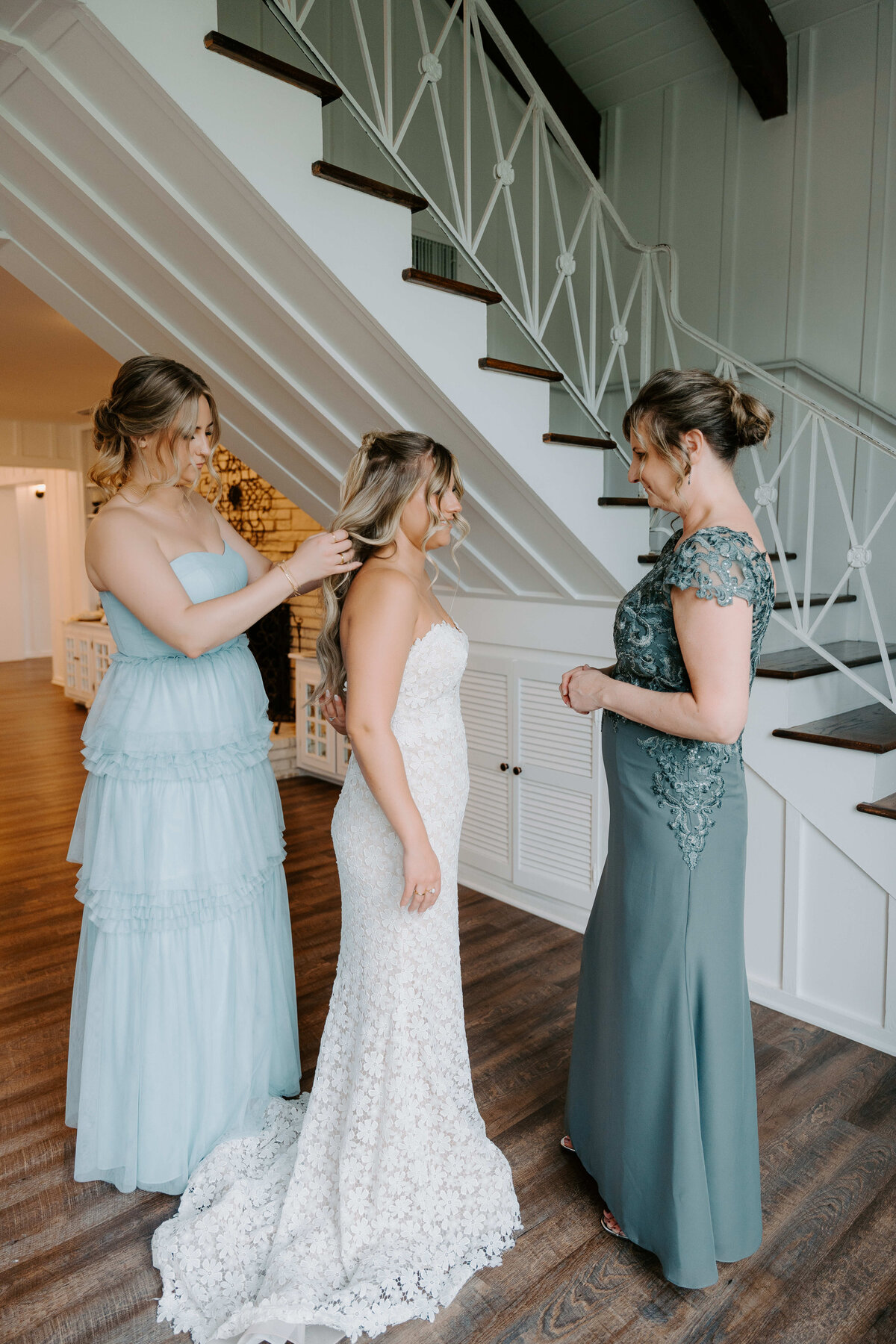 A bride in a white lace gown stands beside a staircase, smiling warmly. Two women in teal and green dresses adjust her hair, conveying a joyful, intimate moment.