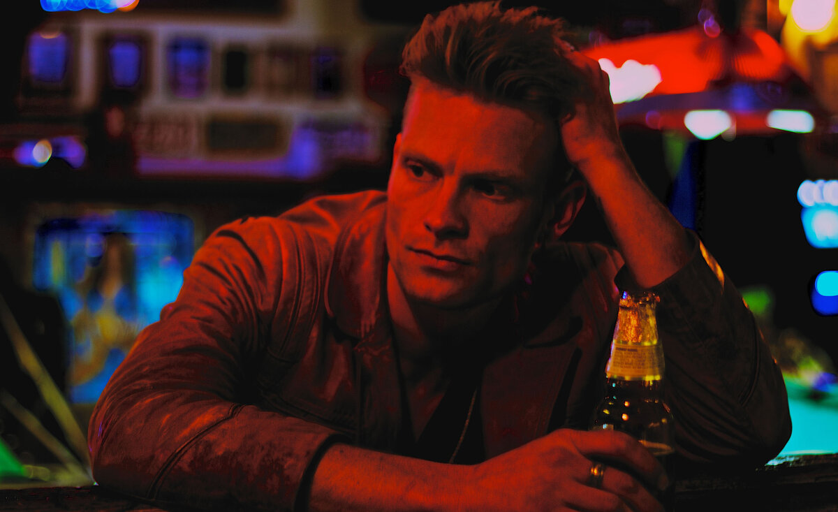 Male musician portrait Ryan Guldemond sitting against bar holding beer with red light cast over face