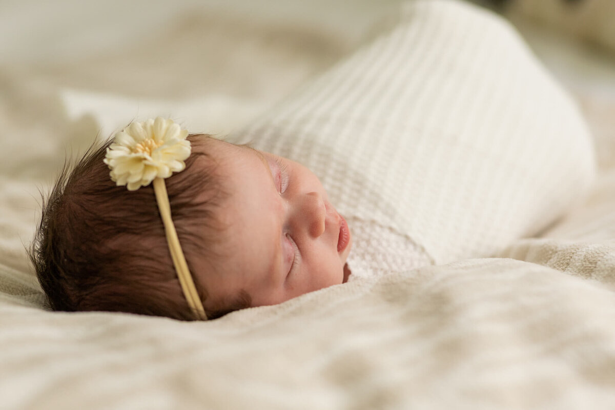 newborn baby girl in a cream wrap and flower bow laying on the bed
