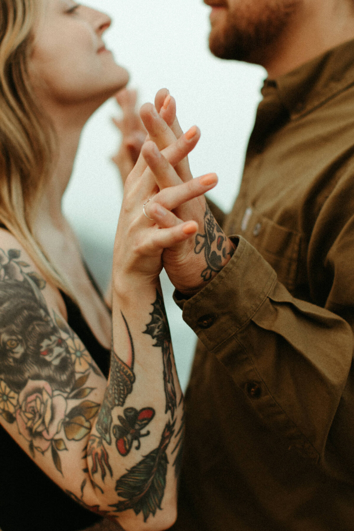 A guy and a girl with tattoos stand facing each other with their fingers intertwined.