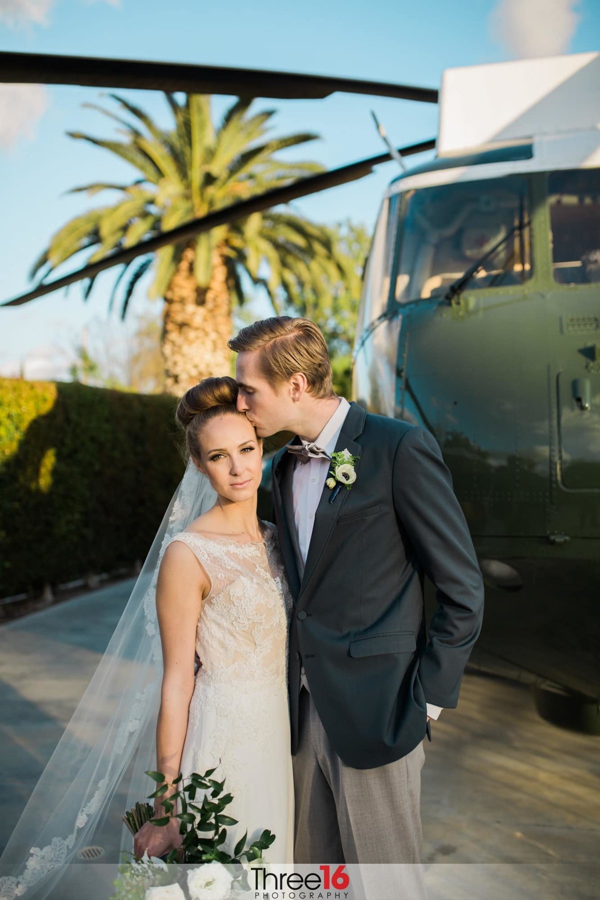 Groom kisses the side of his Bride's head as he embraces her from the side as they stand in front of the Army One helicopter on the grounds of the Richard Nixon Library in Yorba Linda