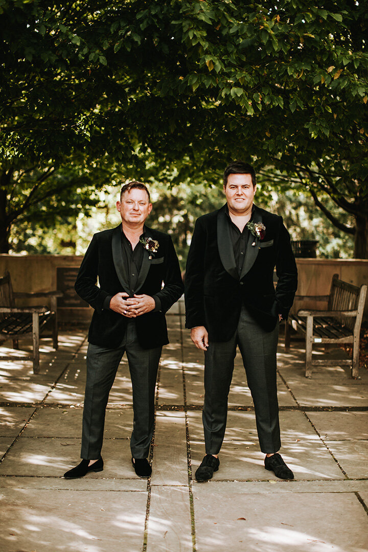 Two grooms wearing black tuxedos pose side by side in an outdoor quart yard.