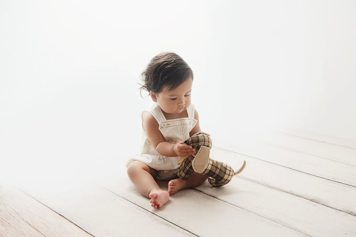 A young toddler in white overalls plays with a stuffed dog on the floor of a studio