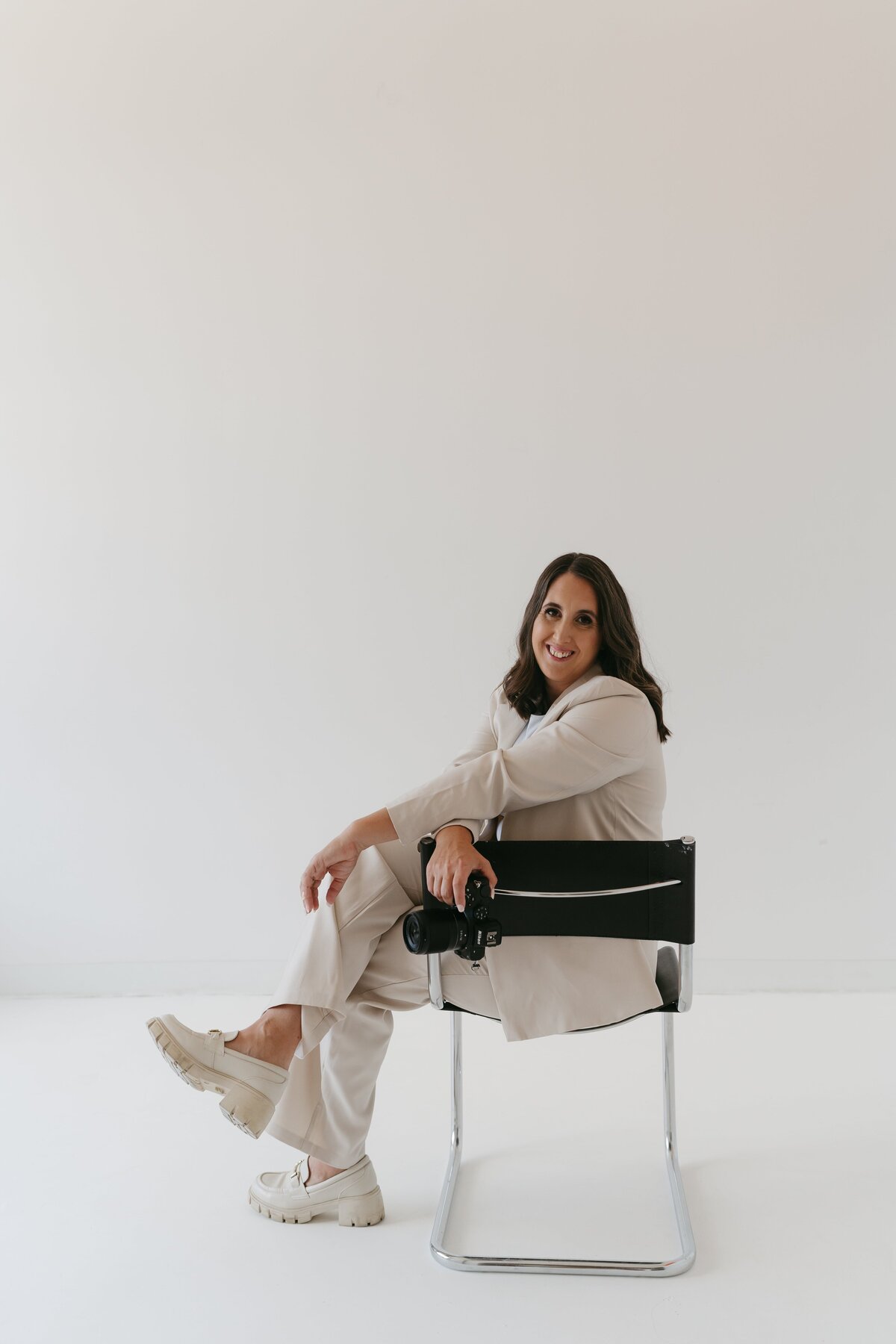 A smiling woman in a beige suit sits on a modern chair against a white background, holding a camera, conveying a professional and confident demeanor.