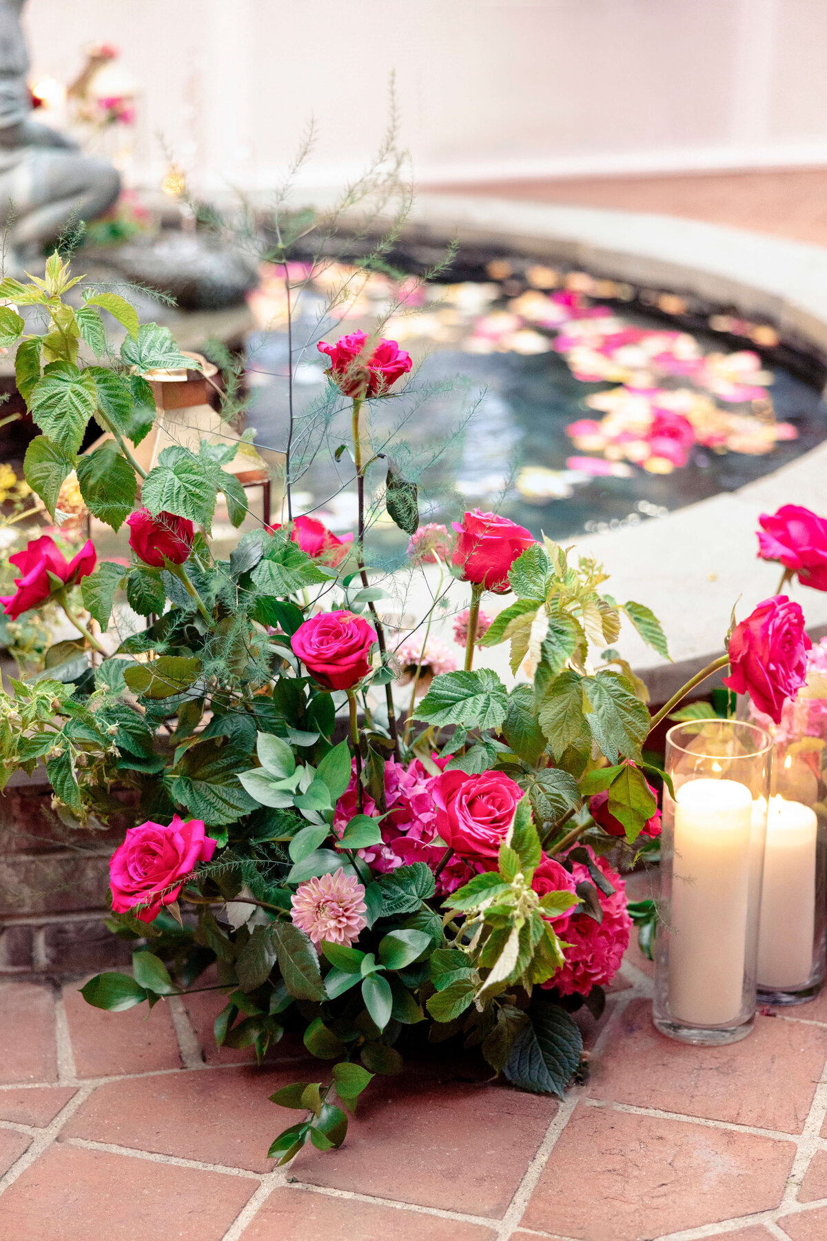 A lush arrangement of pink and red roses with greenery is placed by a decorative fountain. The water is dotted with floating petals, and three tall lit candles stand nearby, creating an elegant and romantic ambiance.