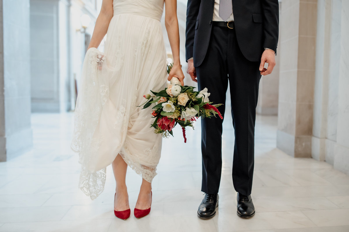 Wedding couple at San Francisco City Hall.  Detail photo from waist down. Bride holds white dress and flowers wearing red shoes. Groom wearing black suit.