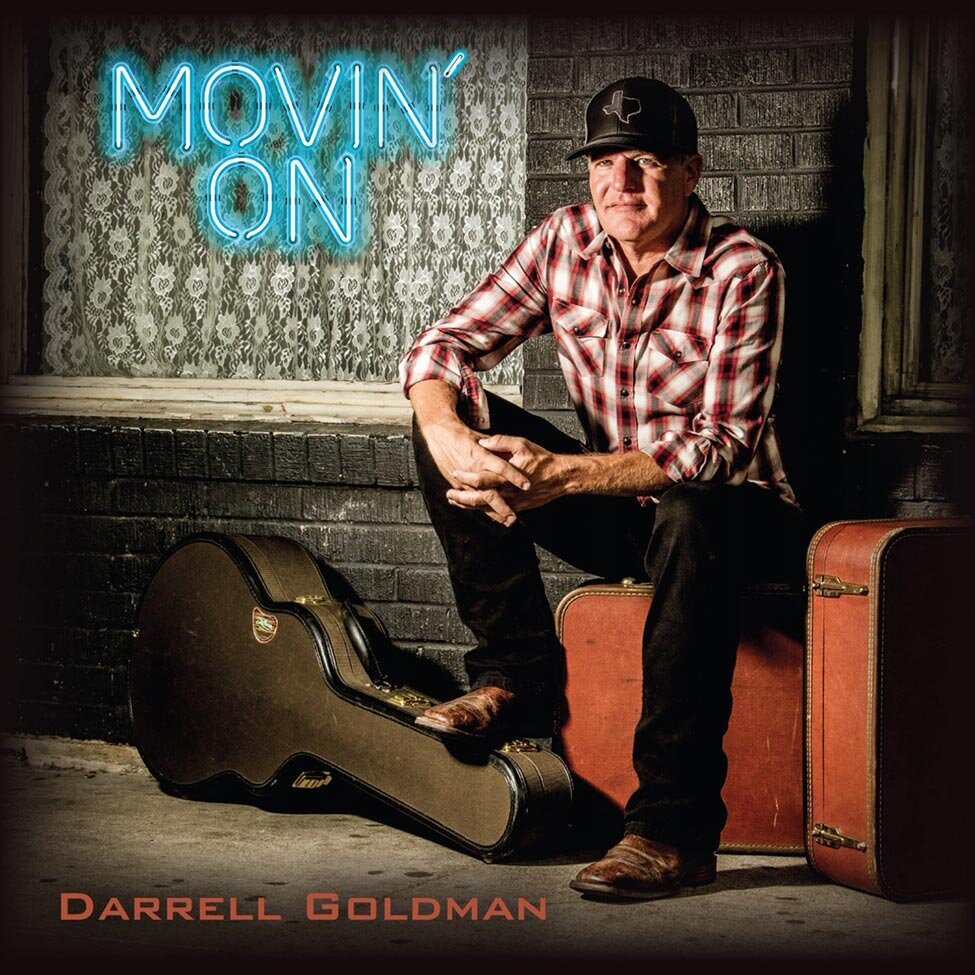Country Music Single Cover Title Movin On Artist Darrell Goldman sitting  on red suitcases wearing plaid shirt and base ball cap foot resting on guitar case in front of him