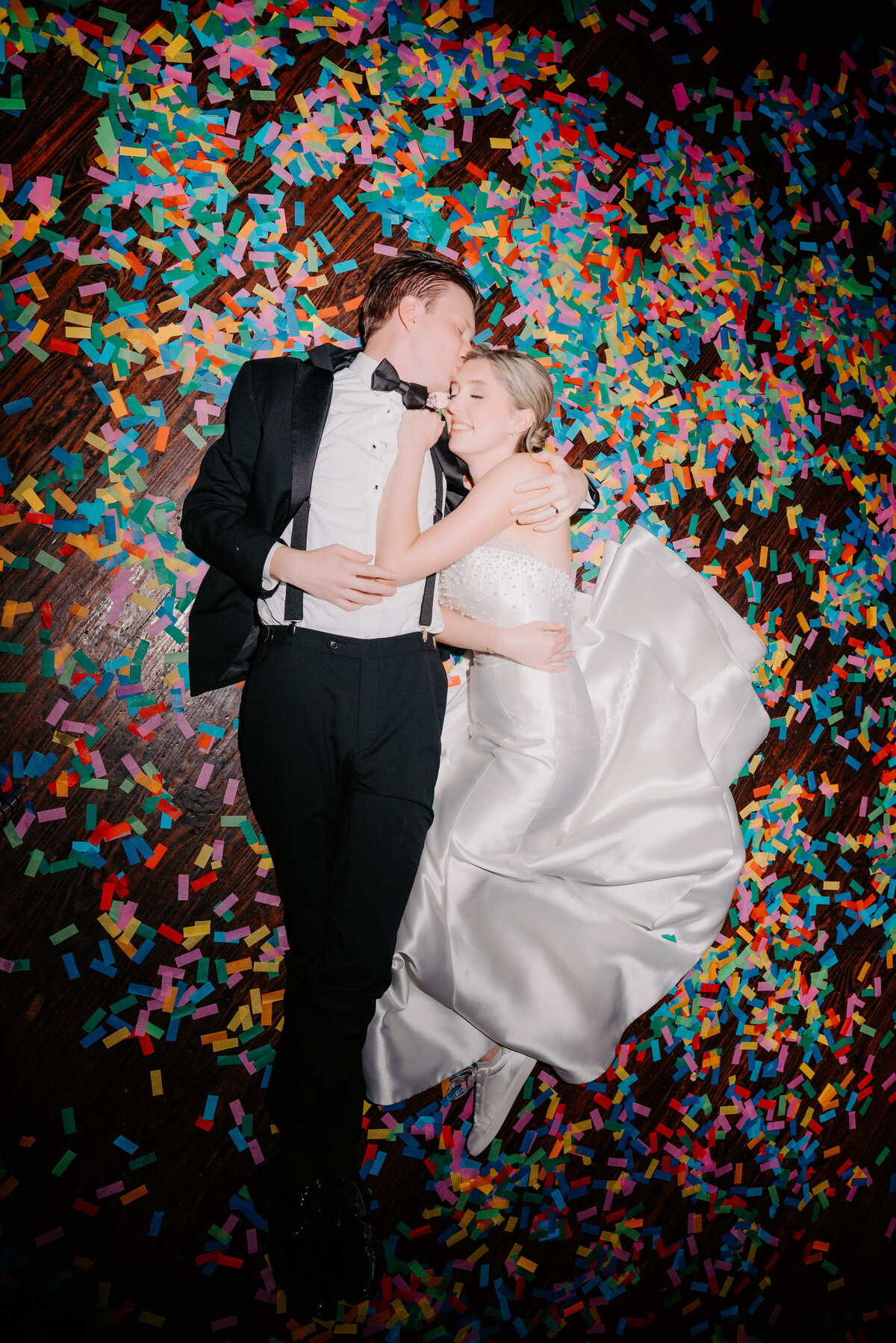 Bride and groom lying on the dance floor surrounded by colorful confetti, embracing and smiling after their wedding celebration