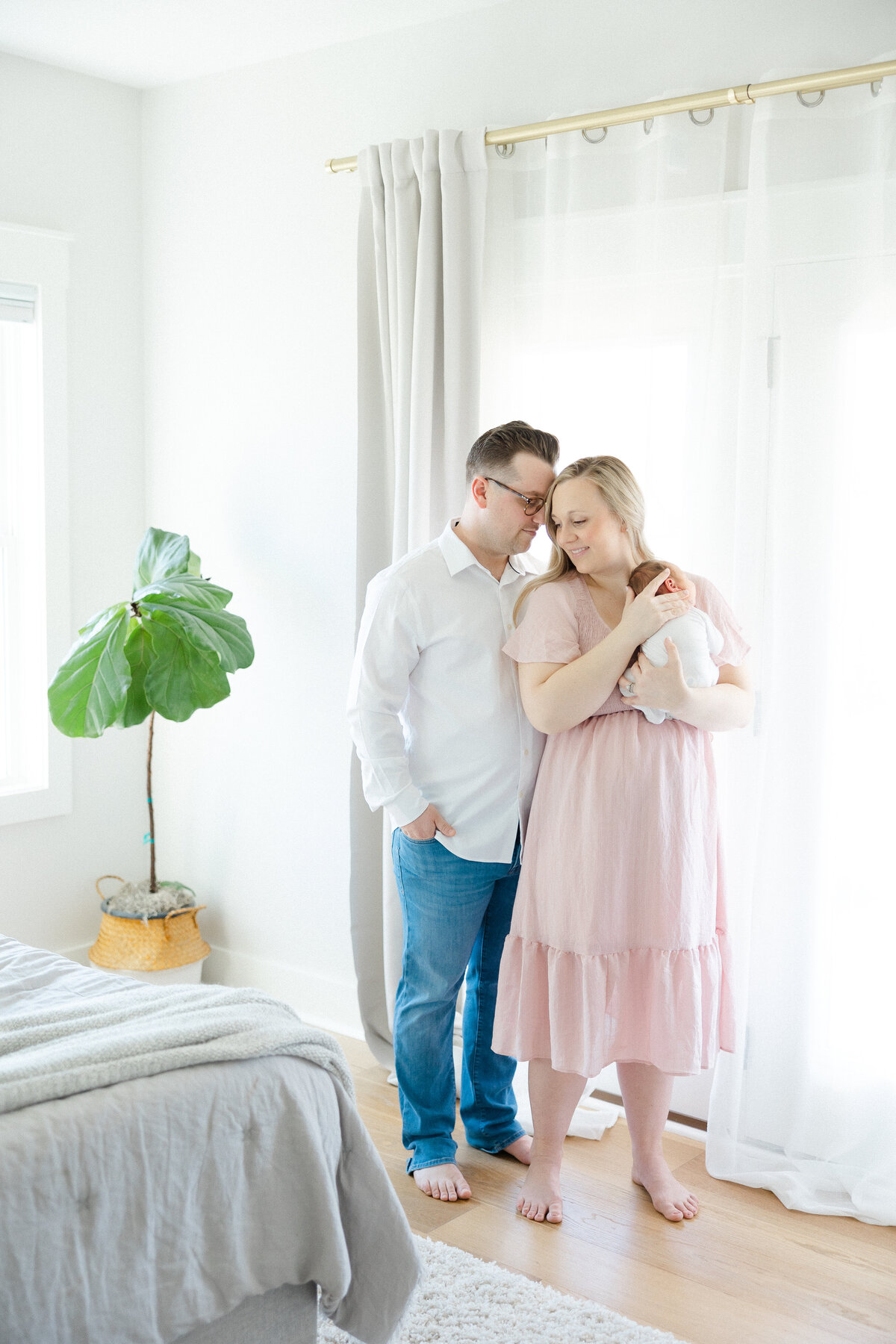 husband and wife standing in front of window holding baby
