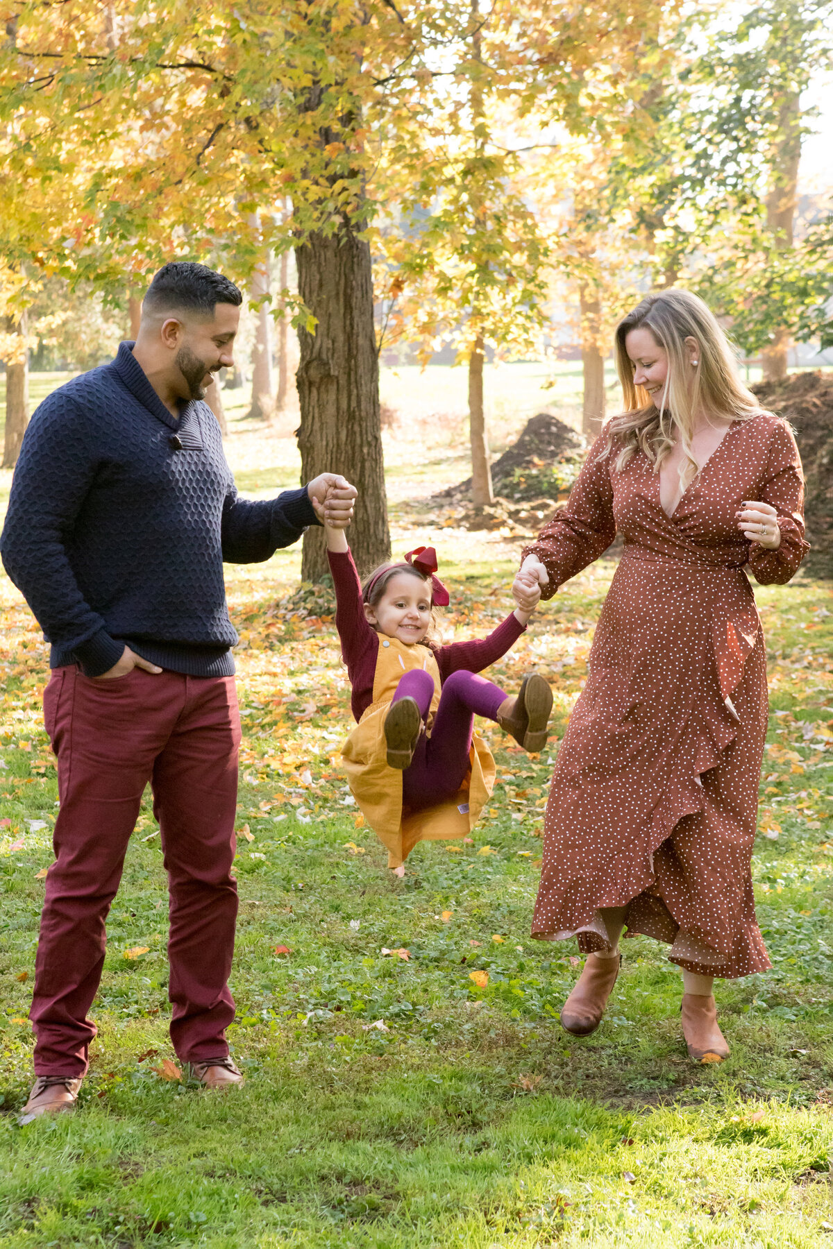 A mother and father swing their young daughter between them in a park with yellow autumn leaves fall mini-session photographer