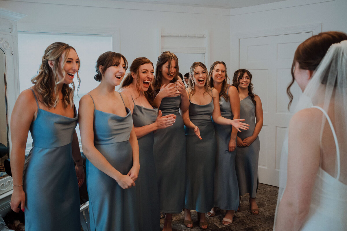 Group of seven bridesmaids in blue satin dresses reacting to seeing bride for the first time