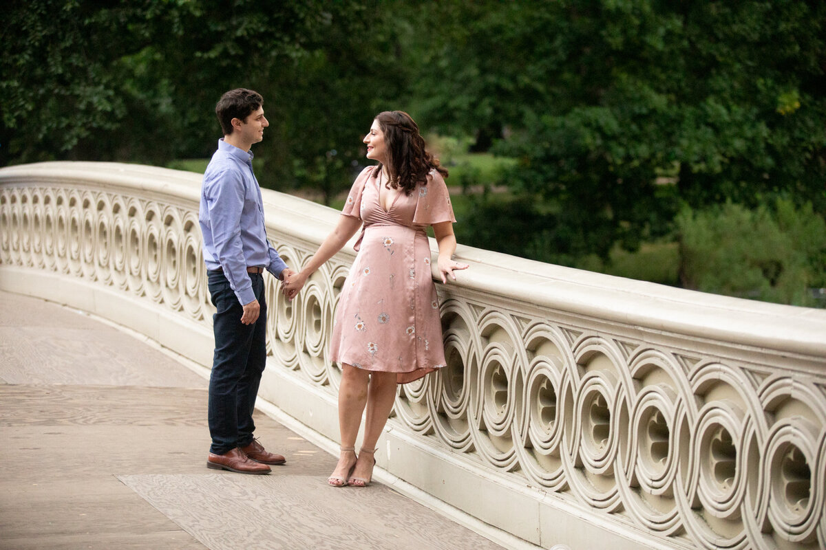 a white pan in blue shirt and black pants holds hands with a woman in a pink dress on bow bridge Central Park engagement session New York wedding photographer