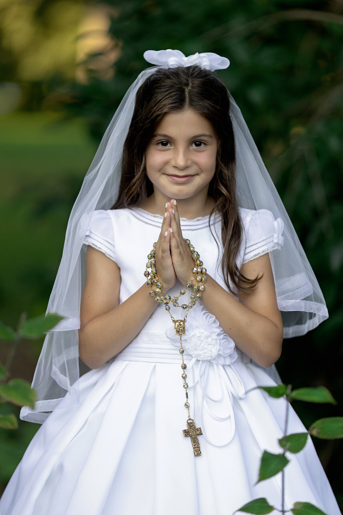 A young girl in a silk white communion dress prays with ornate rosary beads in a park