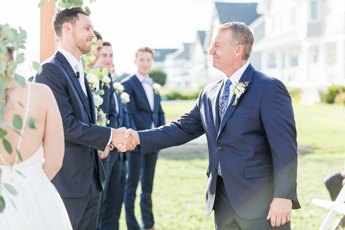 Groom is shaking hands with the father of the bride at a Madison Beach Hotel wedding ceremony. The bride is seen in the foreground. Captured by best New England wedding photographer Lia Rose Weddings.