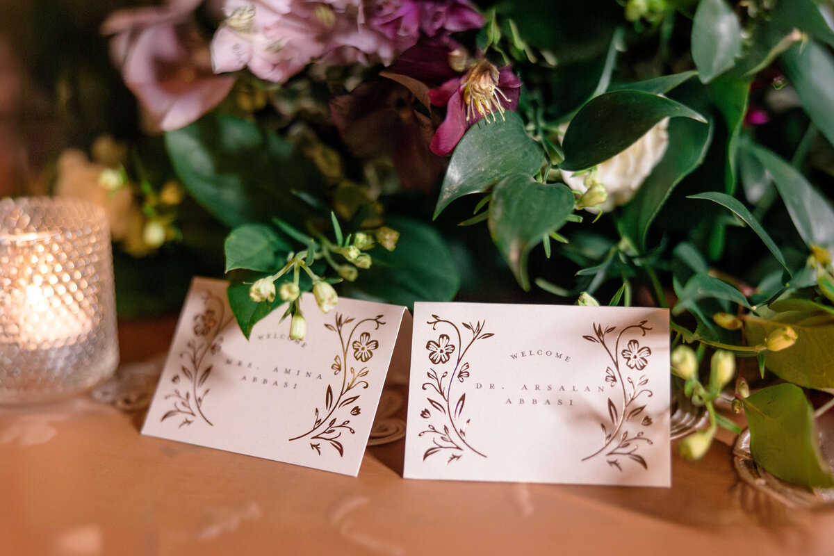 Close-up of two elegant place cards on a table, surrounded by lush greenery and flowers. A small lit candle in a textured glass holder is on the left, adding a warm glow to the scene. The cards feature intricate floral designs and text.