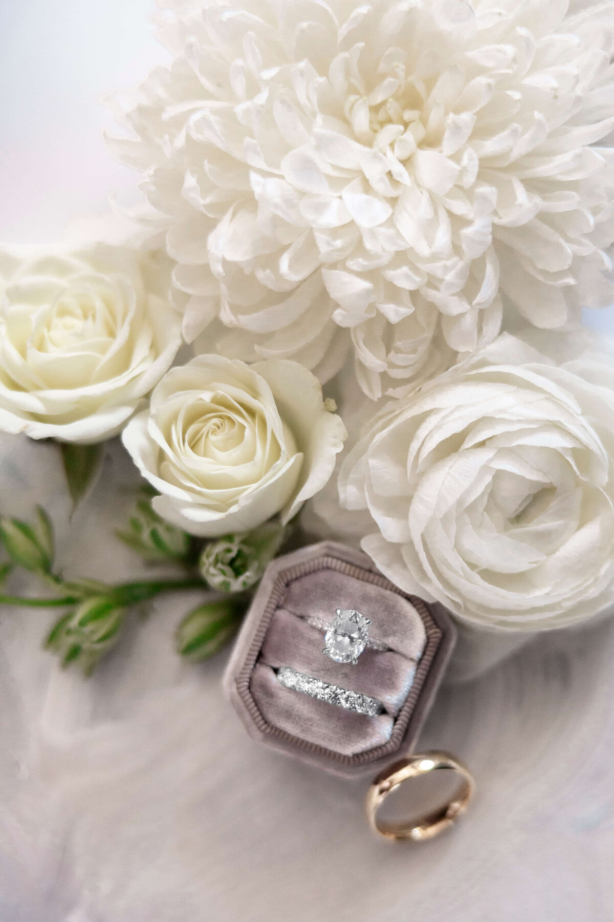 A close-up of a wedding ring set in a gray velvet box surrounded by white roses and a large white flower. The set includes a diamond engagement ring and a wedding band. A separate gold band lies nearby.