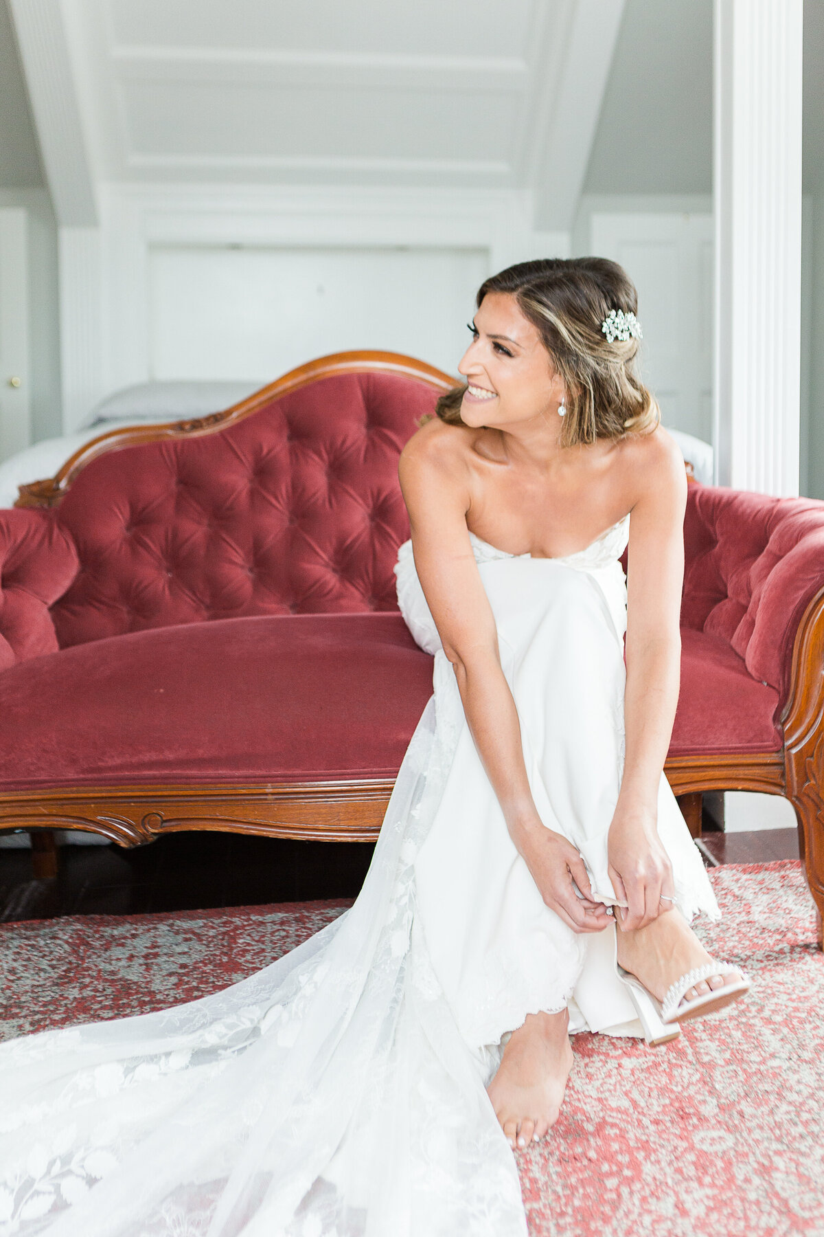 Bride puts on her shows as she is sitting on a burgundy vintage couch before her Five Bridge Inn wedding ceremony. Captured by best Massachusetts wedding photographer Lia Rose Weddings.