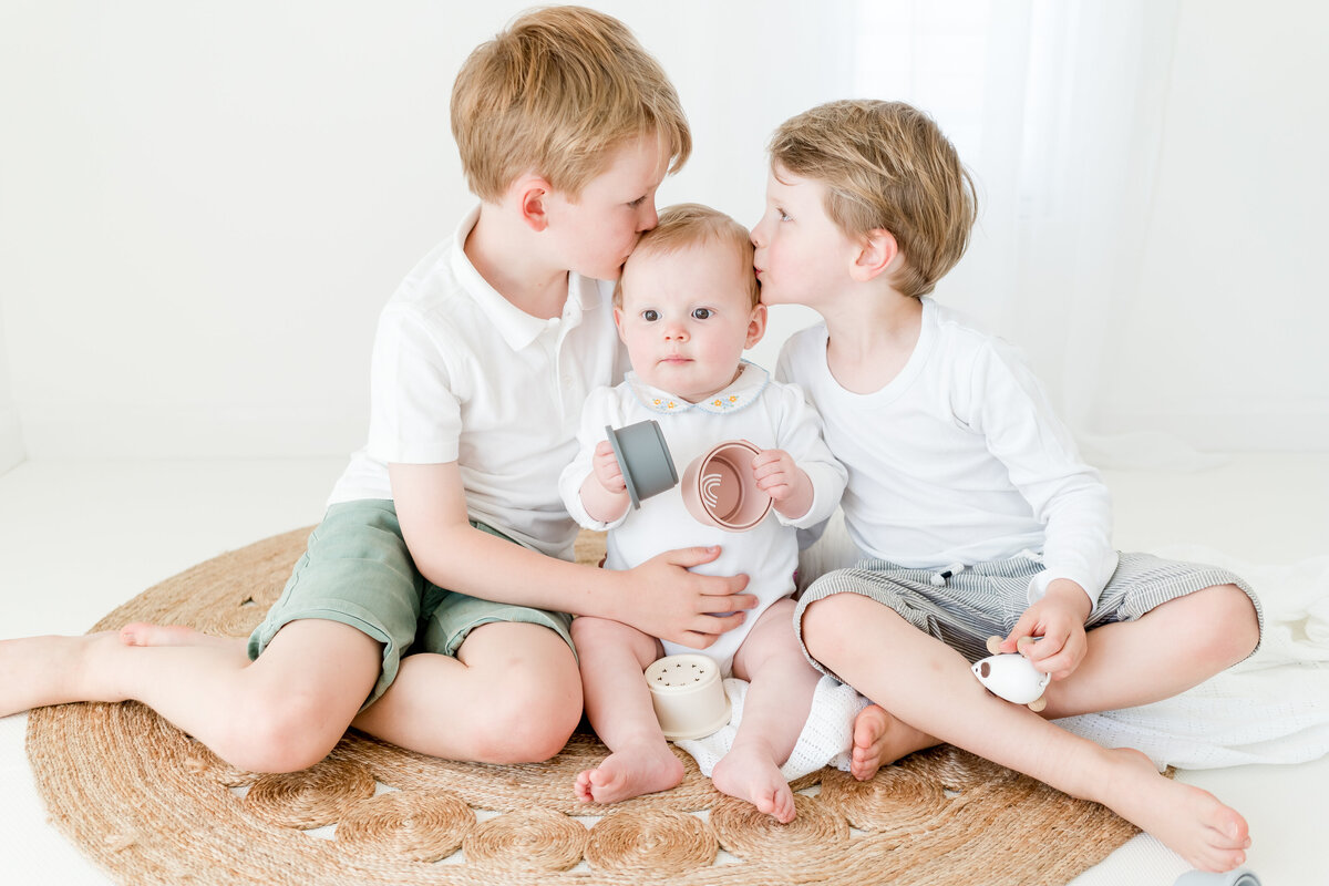 brothers kiss their baby sister on the head during family photo shoot in a studio