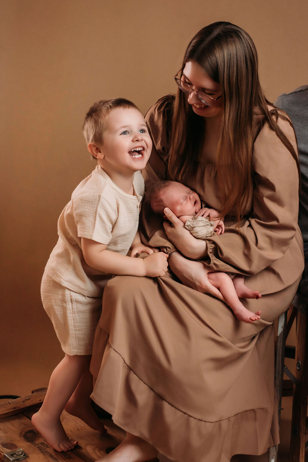A family sits in front of a neutral-toned backdrop––a mother in a tan boho dress holding a newborn baby in a beige outfit while a young child in a beige outfit dotes over his sister with a smile, captured by a photographer in Bemidji, Minnesota.